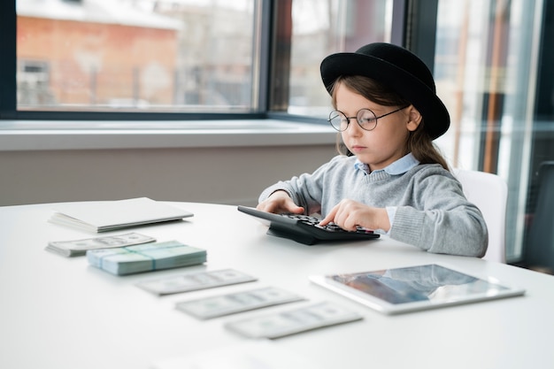 Mały, poważny księgowy w kapeluszu i okularach, korzystający z kalkulatora podczas liczenia pensji kierowników biur przy biurku