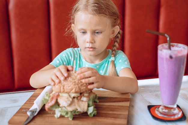 Mały skupiony dzieciak o blond włosach szuka z apetytem na soczystego ogromnego burgera w kawiarni. Portret.