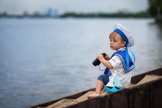 Mały słodki chłopczyk w marynarskim kapeluszu siedzi na brzegu, patrzy przez lornetkę i bawi się łódką