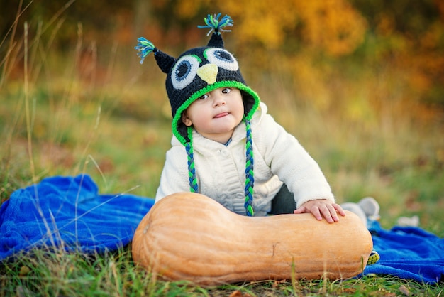 Mały śmieszny chłopiec w czapka z dzianiny pokazuje język, siedząc na trawie z dyni i jesienne drzewa