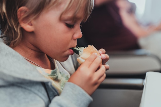 Mały, szczery chłopczyk w wieku pięciu lat je burgera lub kanapkę siedząc w samolocie w samolocie lecąc z lotniska dzieci gryzą dziecko w samolocie jedząc obiad lub kolację