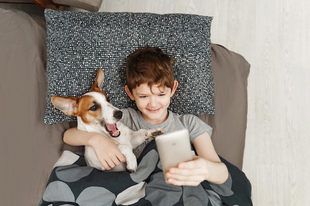 Mały szczęśliwy chłopiec leży na łóżku z psem jack russell terrier i robi sobie selfie.