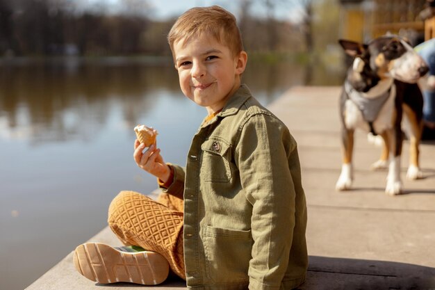 Mały uroczy chłopiec siedzi na zewnątrz i je lody Woda w jeziorze i słoneczna pogoda Dziecko i słodycze Cukier Dzieciak cieszą się pysznym deserem Dziecko w wieku przedszkolnym w swobodnym ubraniu Pozytywne emocje