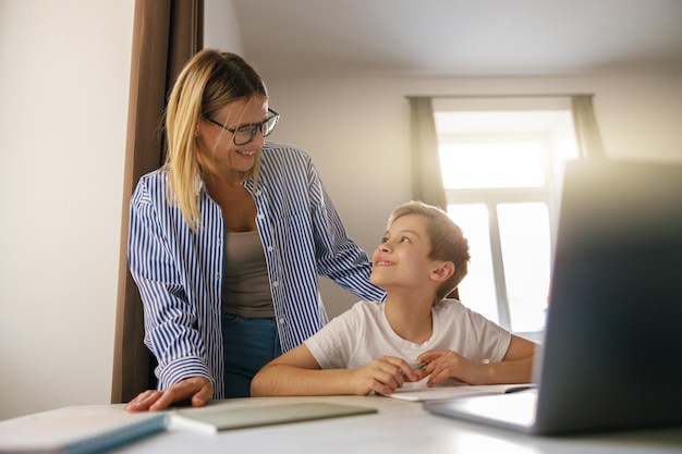 Zdjęcie mały uśmiechnięty uczeń korzystający z laptopa do odrabiania lekcji w szkole z pomocą swojej mamy