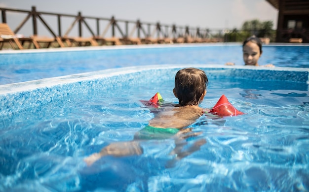 Mały wesoły zabawny dzieciak w jasnych, nadmuchiwanych rękawach pływa w płytkim basenie dla dzieci i bawi się w chowanego ze swoją starszą siostrą podczas słonecznego, ciepłego urlopu