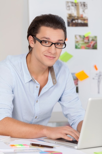 Mam tyle pomysłów. Przystojny młody mężczyzna w okularach pracujący na komputerze i uśmiechający się do kamery siedząc w swoim miejscu pracy