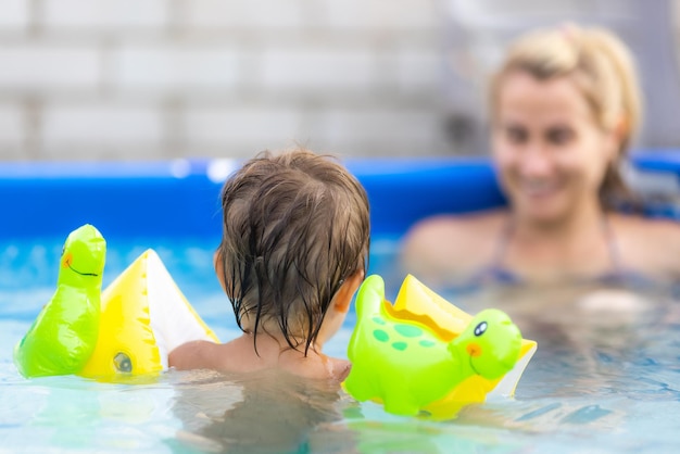 Mama bawi się z nagim dzieckiem w zarękawkach w basenie na tle letniego zachodu słońca