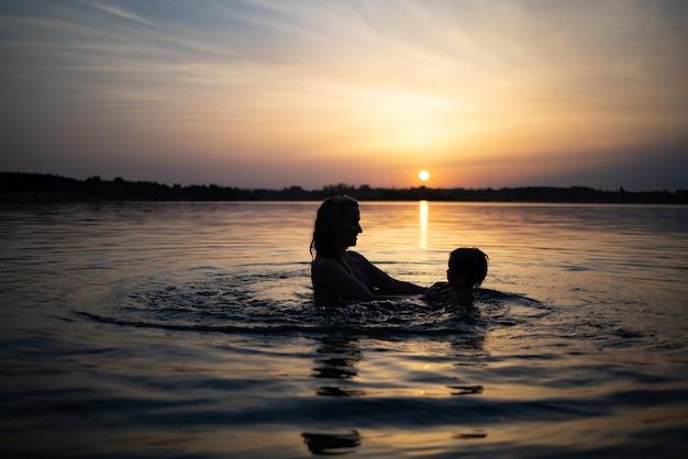 Mama bawi się z nagim dzieckiem w zarękawkach w jeziorze na tle letniego zachodu słońca