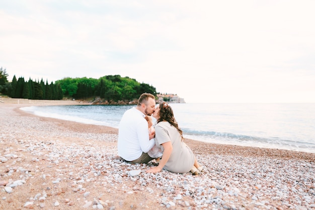 Mama całuje tatę z małą dziewczynką w ramionach siedząc na plaży w pobliżu willi milocer na