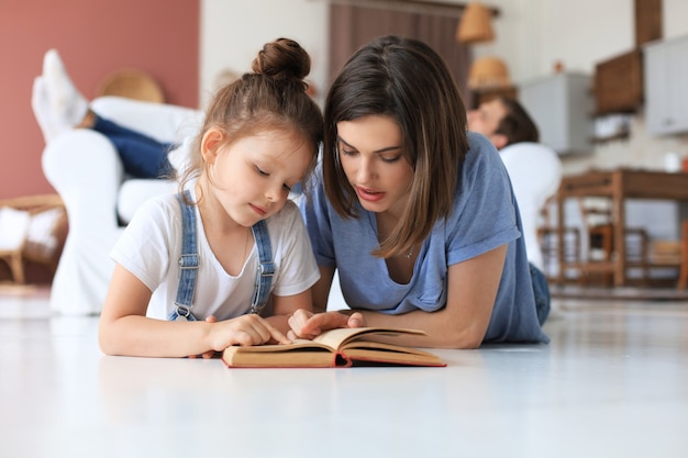 Mama i córeczka czytają razem książkę, leżąc na podłodze w salonie.