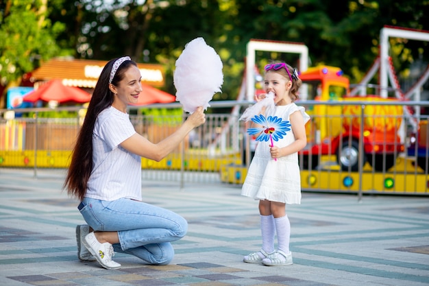 Mama i córeczka jedzą watę cukrową i uśmiechają się ze szczęścia w wesołym miasteczku w lecie