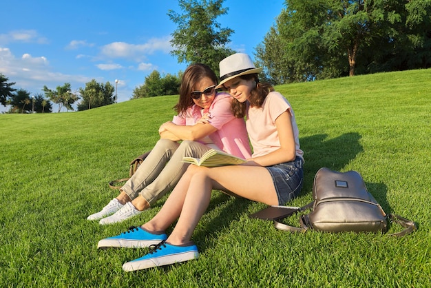 Mama i córka nastolatka razem siedząca na zielonym trawniku, odpoczywająca, dziewczyna pokazująca mamie książkę, rozmawiający rodzic i dziecko