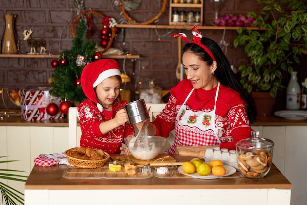 Mama i córka w ciemnej kuchni z choinką przygotowują imbirowe ciasteczka na Nowy Rok lub Boże Narodzenie, uśmiechając się i bawiąc się razem w oczekiwaniu na święta w czapce Świętego Mikołaja