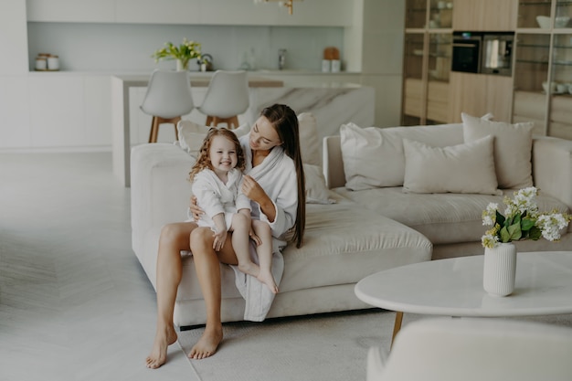 Mama i córka w domowych ubraniach siedzą na wygodnej sofie w salonie.