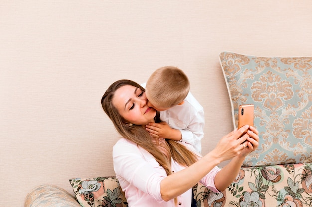 Mama i dziecko robią sobie selfie w przytulnym salonie. mama i syn robią śmieszne miny i robią sobie zdjęcia w aparacie