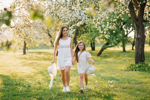 Mama i jej córka w białych sukienkach biegają wiosną w słoneczny dzień po kwitnącym jabłoniowym sadzie i dobrze się bawią