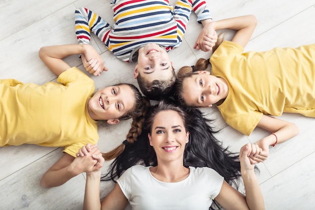 Mama i jej trójka dzieci trzymają się za ręce, leżąc na podłodze, patrząc w kamerę.