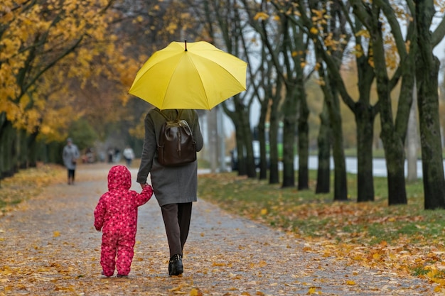 Mama i małe dziecko chodzą pod żółtym parasolem w jesiennym żółtym parku trzymając się za ręce. Widok z tyłu.