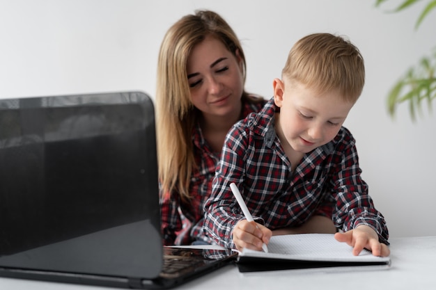 Mama I Syn Odrabiają Lekcje. Student Uczenia Się Na Odległość. Kobieta Pomaga Dziecku W Nauce Tematu Lekcji. Chłopiec Zapisuje Odpowiedzi W Zeszycie. Edukacja Online Poddana Kwarantannie.