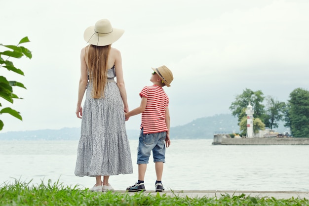 Mama i syn stojący na molo. Morze w tle, latarnia morska i góry w oddali. Widok z tyłu