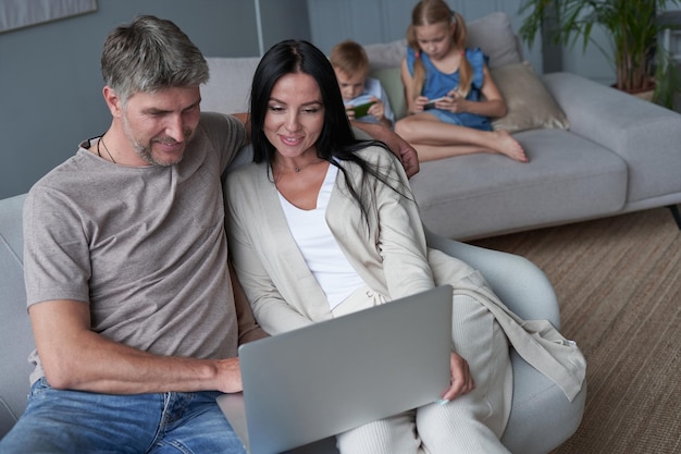 Mama i tata korzystają z laptopa, podczas gdy ich dzieci odpoczywają na kanapie