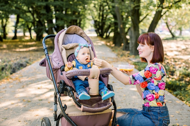 Mama Karmi Córeczkę łyżeczką. Matka Daje Jedzenie Dziesięciomiesięcznemu Dziecku Chodząc Z Wózkiem W Parku. Jedzenie Dla Dzieci.