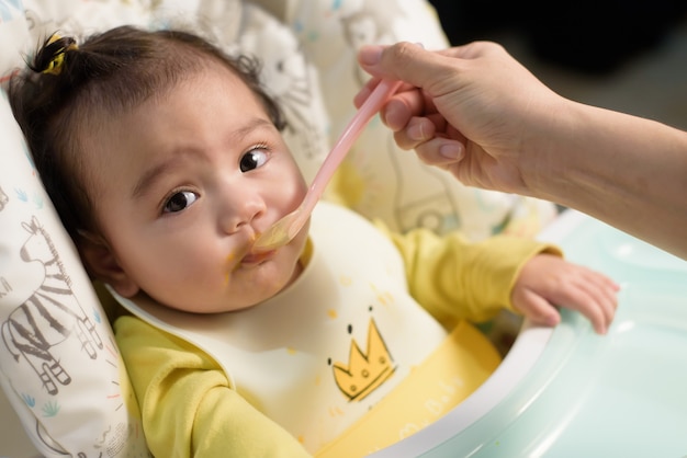 Zdjęcie mama karmi jedzenie swoją małą azjatycką córeczkę łyżką w domu, dziecko patrzy na kamerę