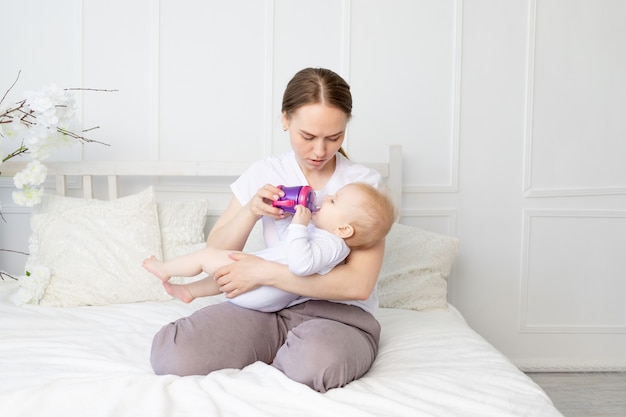 Mama pije lub butelką karmi dziecko na białym łóżku w domu, koncepcja żywności dla niemowląt.