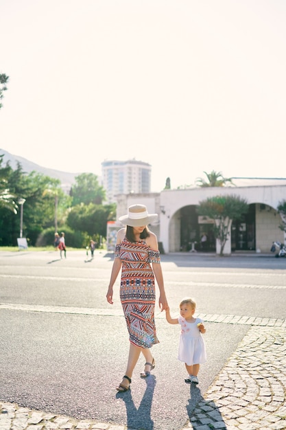 Mama prowadzi za rękę małą dziewczynkę przez plac na tle budynków