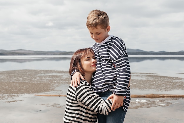 Mama przytula syna na brzegu rzeki. Piękna, nowoczesna mama z szczęśliwym synkiem w pasiastych kamizelkach.