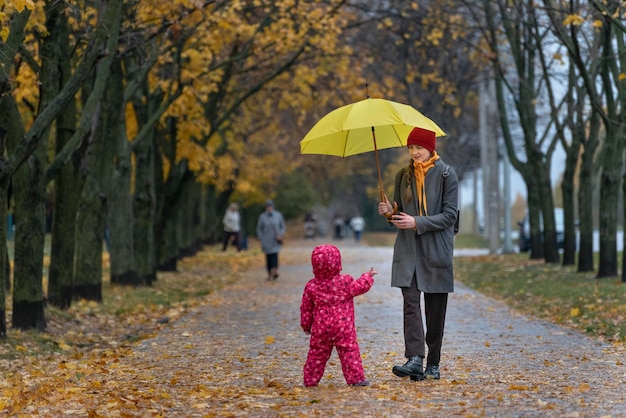 Mama spaceruje z małym dzieckiem w jesiennym parku pod żółtym parasolem