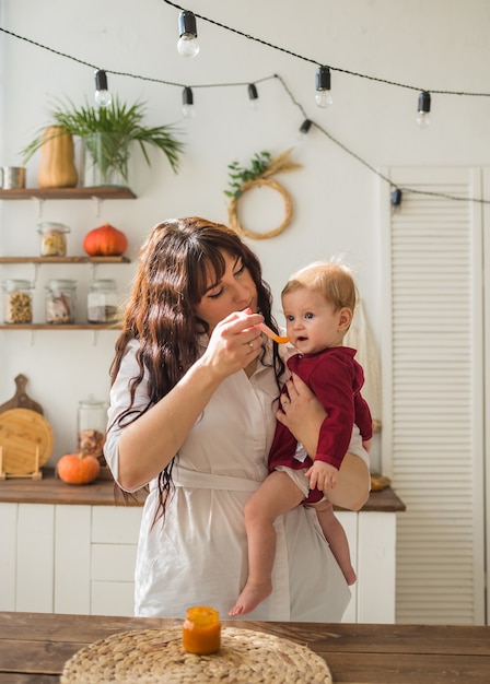 Mama trzyma córeczkę i karmi w kuchni puree z marchwi