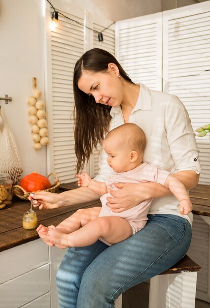 Mama Trzyma Córkę W Ramionach I Karmi łyżką Tłuczone Ziemniaki Na Krześle W Kuchni