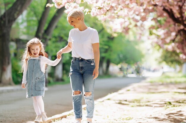 Mama trzyma córkę za rękę na wiosennej ulicy miasta pod gałęziami kwitnącej różowej sakury