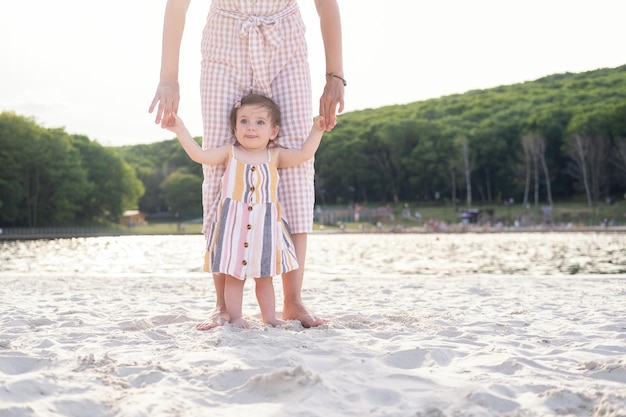 mama uczy córkę chodzić trzymając się za ręce po plaży na piasku w słoneczny dzień