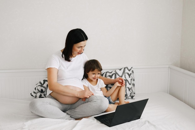 Mama w ciąży z córeczką siedzącą na łóżku przy laptopie