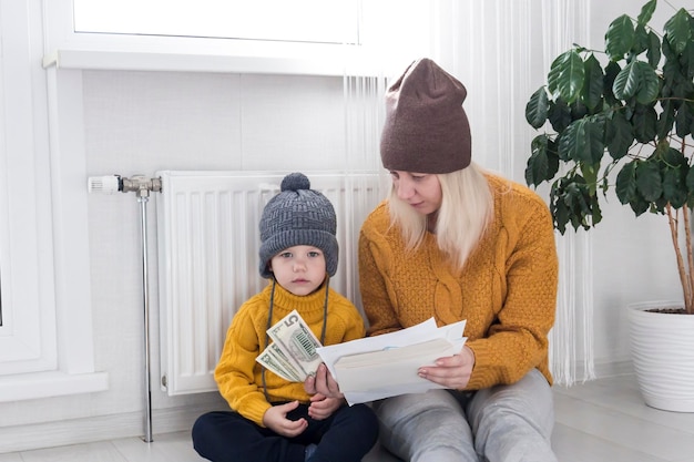 Mama z małym dzieckiem w żółtym swetrze i czapeczkach liczy pieniądze i myśli jak zapłacić rachunki i podatki przy grzejniku z termostatem