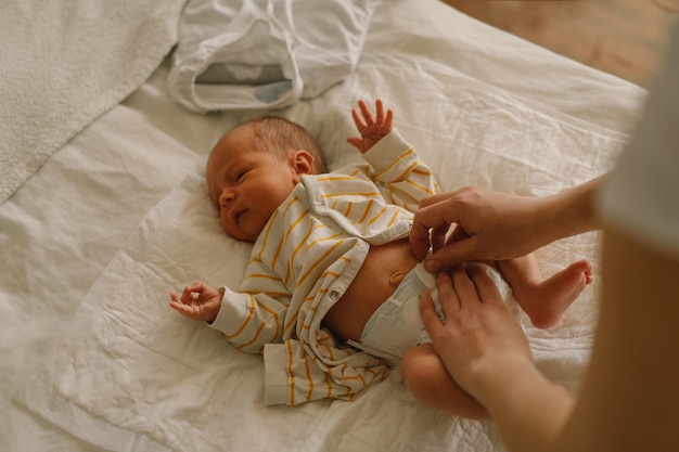 Mama zmienia pieluchę noworodka. Szczęśliwa młoda matka bawi się z dzieckiem podczas zmiany pieluchy na łóżku. Szczęśliwe macierzyństwo