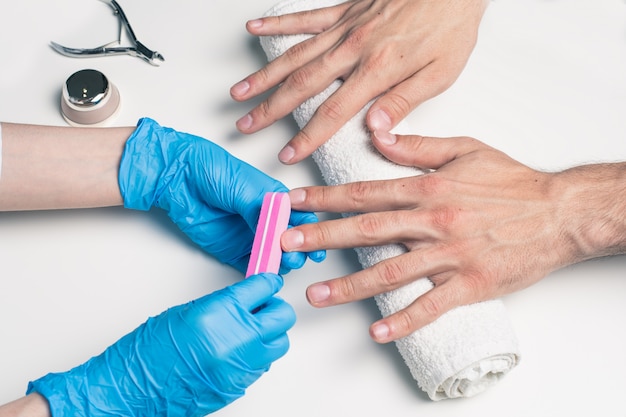 Manicure męski. Kosmetolog w gumowych rękawicach pilnikuje paznokcie na męskich dłoniach.