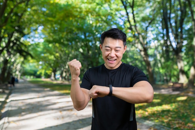 Maraton młody azjatycki sportowiec stoi w parku w sportowym ubraniu i patrzy na bransoletkę fitness