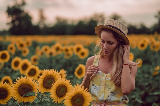 Marzy młoda kobieta w żółtej sukience i kapeluszu, trzymając włosy rękami w polu słoneczników w lecie, widok z przodu. Patrząc w dół. skopiuj miejsce