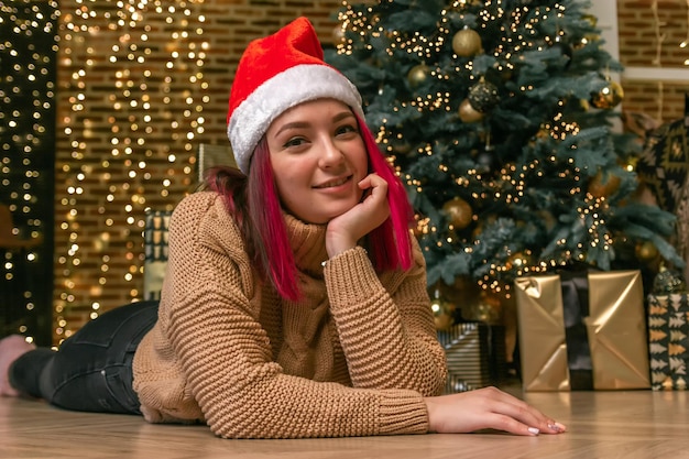 Marzycielska urocza młoda dama w swetrze i czapce świątecznej na podłodze na tle choinki z prezentami