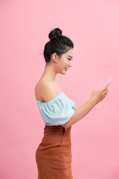 Marzycielski azjatycki piękna młoda kobieta posiada nowoczesne urządzenie elektroniczne, odizolowane na różowym tle.