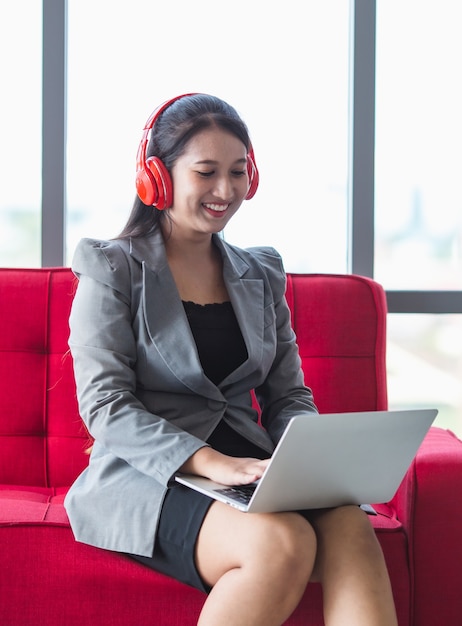 Marzycielski azjatycki przedsiębiorca w słuchawkach siedzący na kanapie w korytarzu centrum biznesowego i cieszący się piosenkami z laptopa notebook z łatwą pozą podczas przerwy w pracy