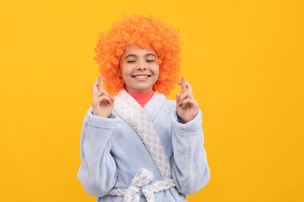 Marzycielski dzieciak w fantazyjnej pomarańczowej peruce nosi szlafrok do domu, życząc ze skrzyżowanymi palcami