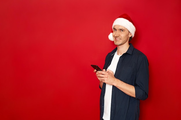 Marzycielski facet w noworocznej czapce z telefonem w dłoni pisze wiadomość, myśli o gratulacjach