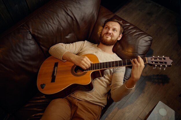 Marzycielski uśmiechnięty, zamyślony, romantyczny mężczyzna leżący na kanapie grający na gitarze, patrzący w górę z góry