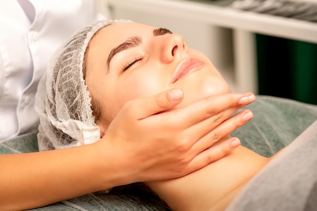 Masaż twarzy Ręce masażysty masujące szyję młodej kobiety rasy kaukaskiej w salonie spa koncepcja masażu zdrowotnego