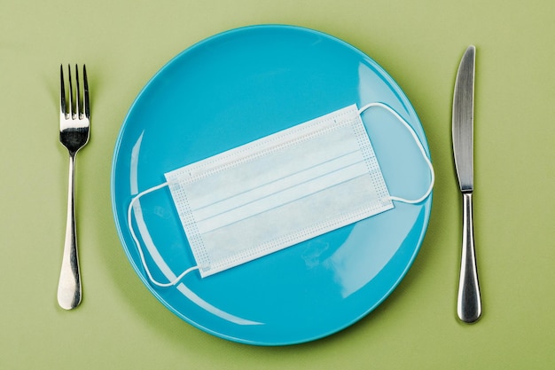 Maska medyczna na pustym talerzu z widelcem i nożem na zielonym tle Pojęcie żywienia i ochrony przed wirusem