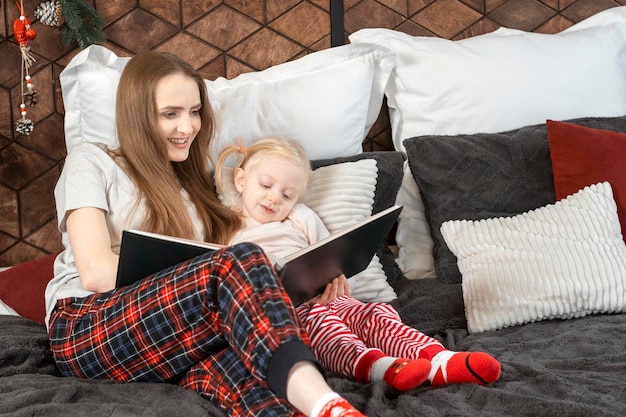 Matka czytająca bajkę do cute blond dziewczyny Matka i córka czyta książkę leżącą w łóżku w pobliżu choinki